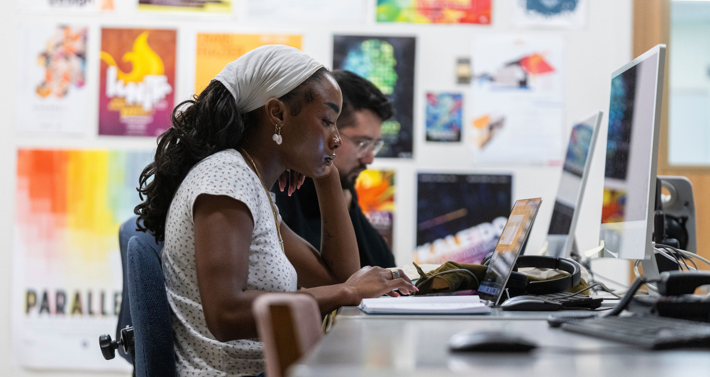 Two students work at computers with design art hanging on the wall in the background.