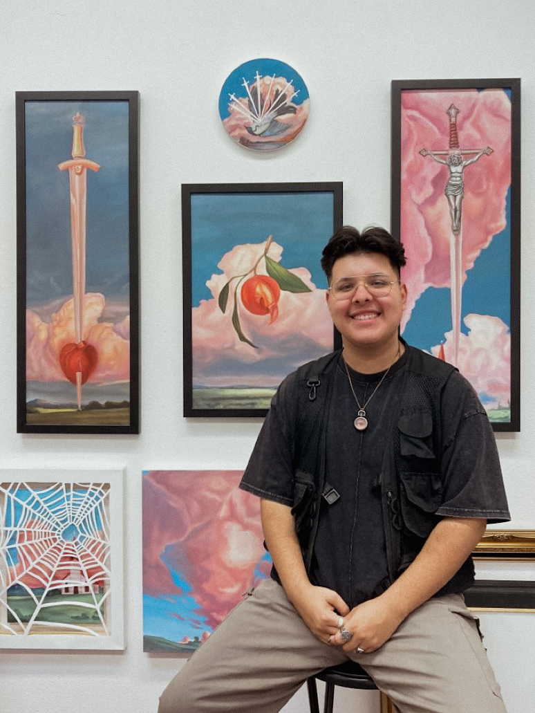 Artist Jose Soria sits on a stool in front of 6 painting that are primarily blue and pink in color