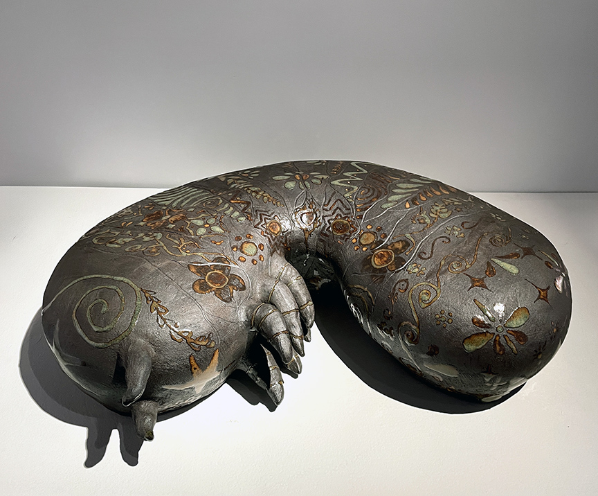 Large grey ceramic grub with rust colored organic shapes and flowers carved into it. The grub is sitting on a white pedistl with a light grey wall behing it. 