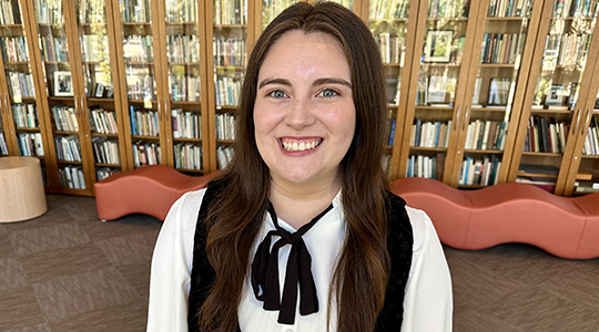 Headshot of Kayla Mendoza, with bookcases full of books in the background.