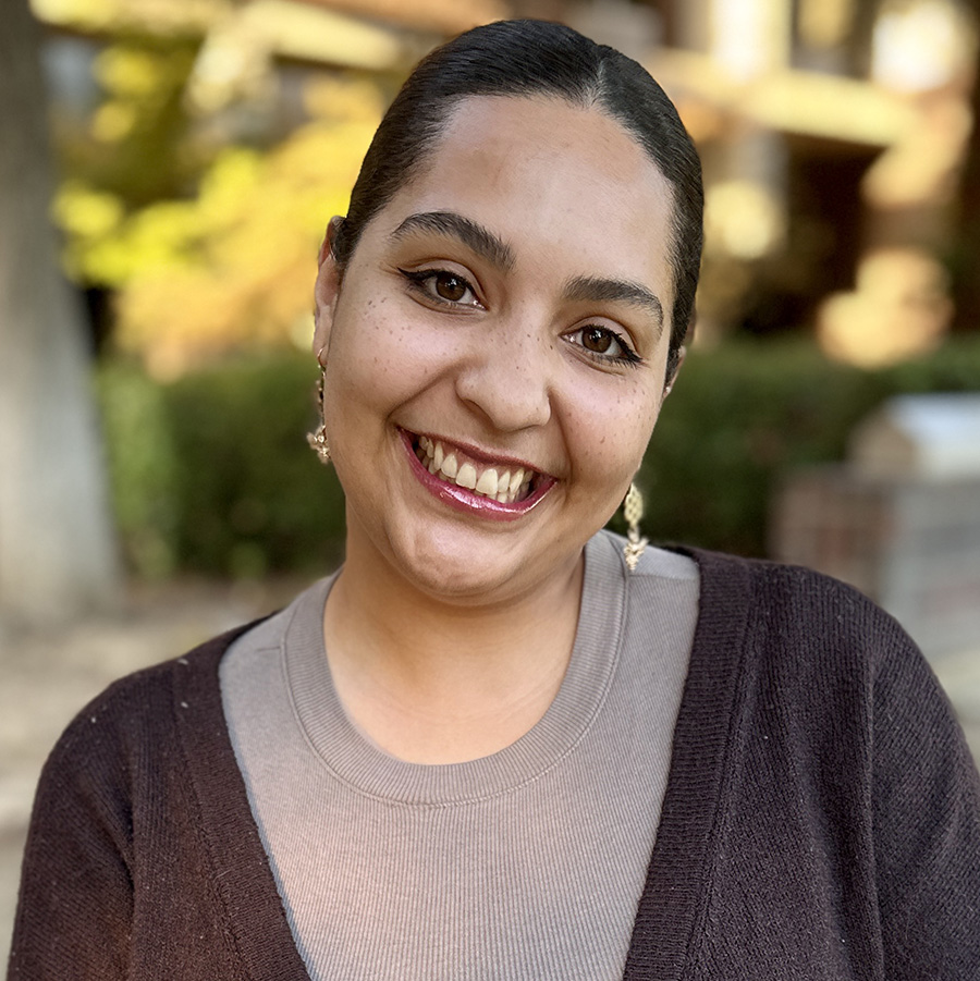 Headshot of Halima, a smiling woman with slick back hair wearing a gray shirt and dark sweater.