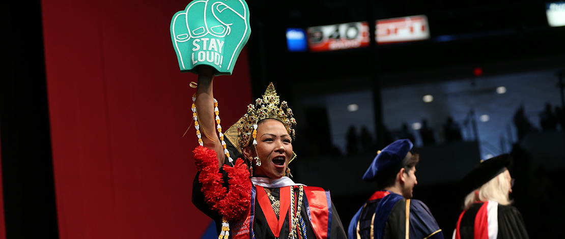 MA graduate Debbie Sayachack holds up a foam fist that reads "Stay Loud" while walking across the stage at commencement.