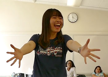 Student stretches out her hands and fingers in exclamation with a big smile as other students look on.