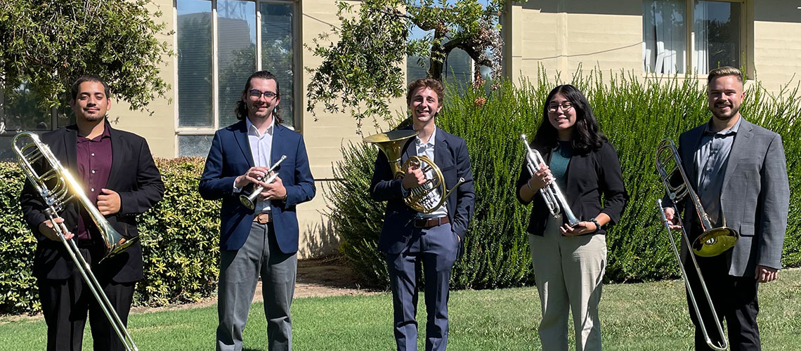 Clendenin Brass Quintet, Erik Nickell, Isley Parada, Carson Kimber, Key Poulan IV and Maynor Quiroa hold their instruments while standing on a green lawn in from of a building with bushes in front.
