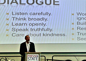 Dr. Andrew Fiala addresses an unseen crowd in front of a large projection image which talks about "dialogue" in bullets are "Listen Carefully, Think Broadly, Learn Openly, Speak Truthfully, Care about Kindness" along with several other bullets that are not fully in the image.